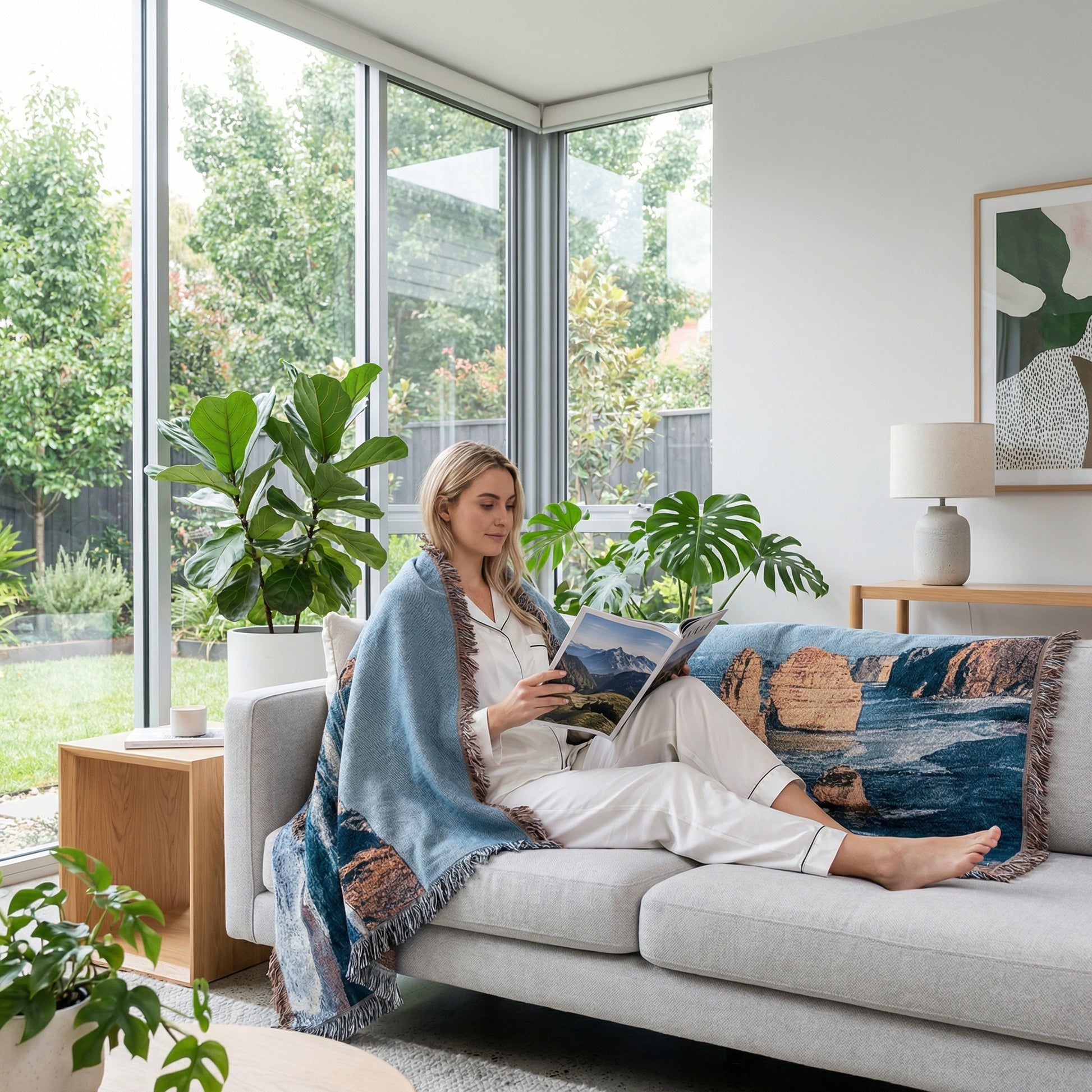 Woman on a gray sofa wrapped in a blue knitted blanket with a natural landscape design, reading a magazine.