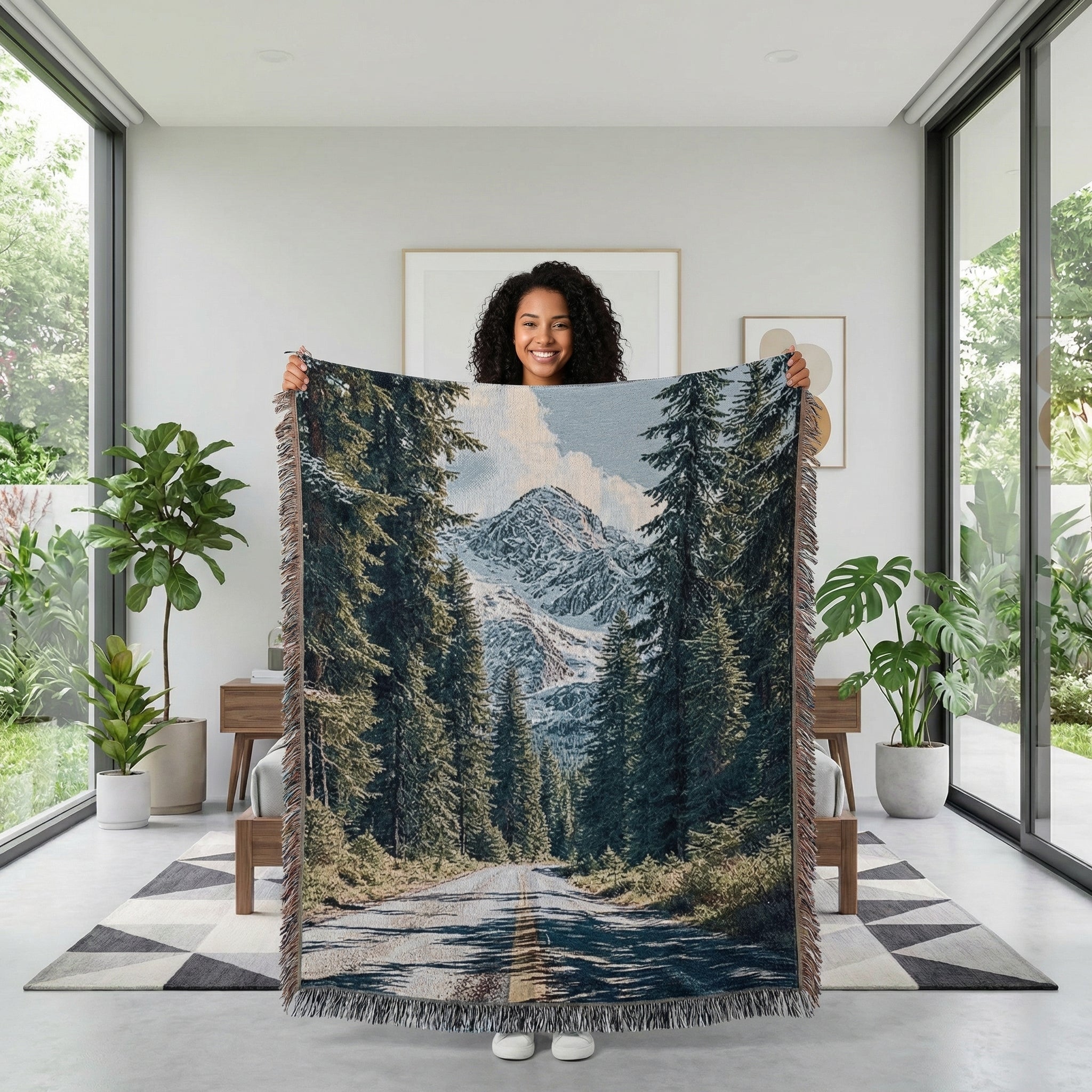 Smiling woman holding a large woven blanket with a natural landscape design of a road, pine trees, and snowy mountains.