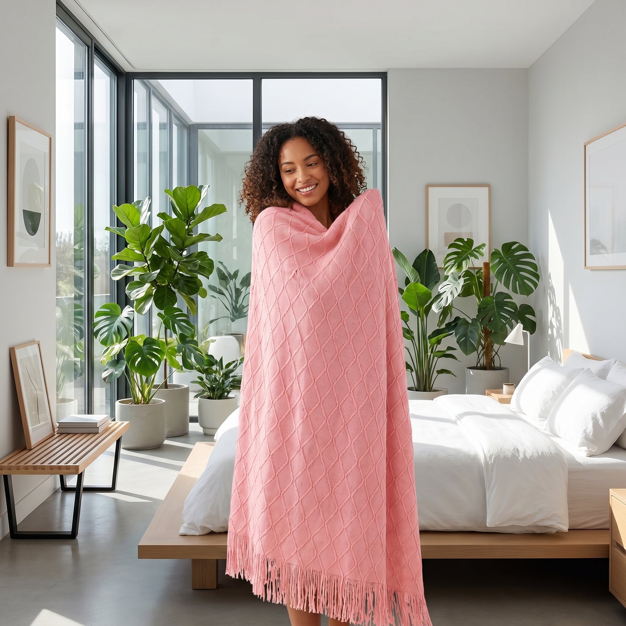 Smiling woman wrapped in a soft pink knit throw blanket with tassels, in a bright bedroom.