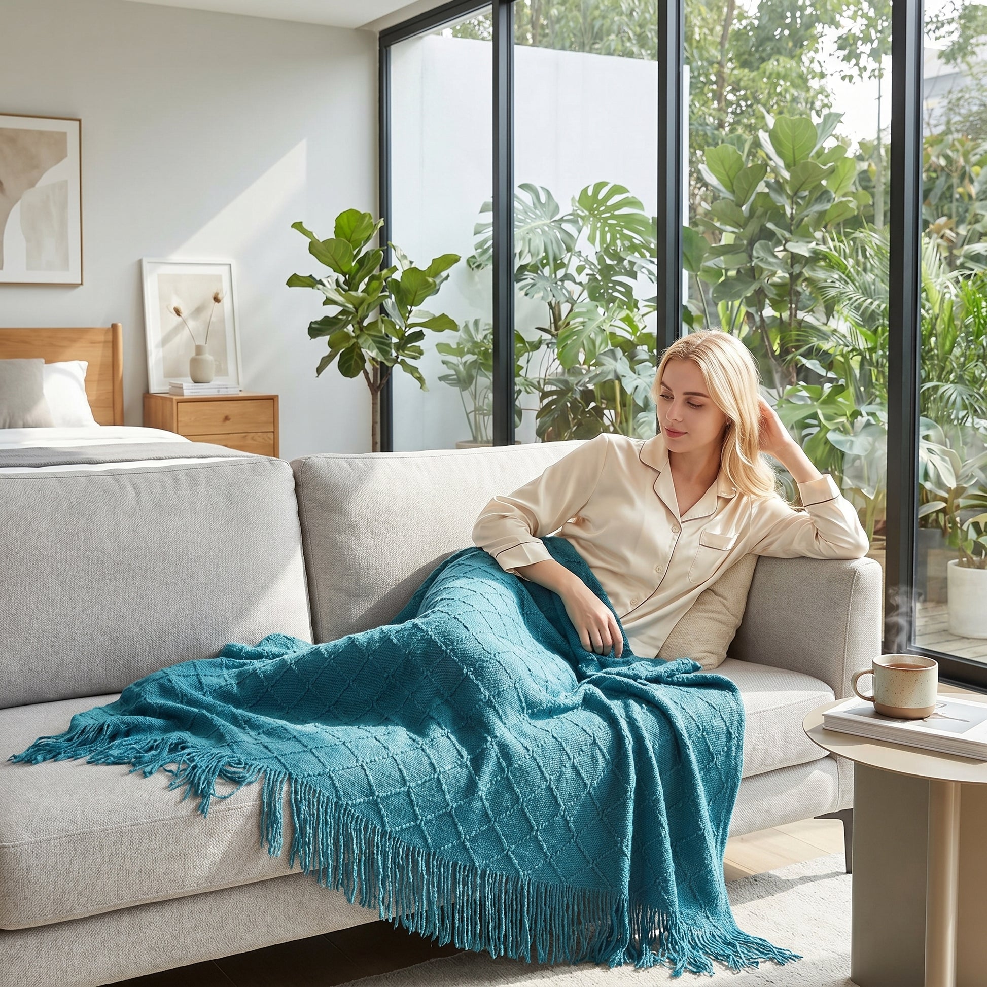 Blonde woman relaxing on a gray sofa, covered by a teal plaid knit throw blanket with tassels.