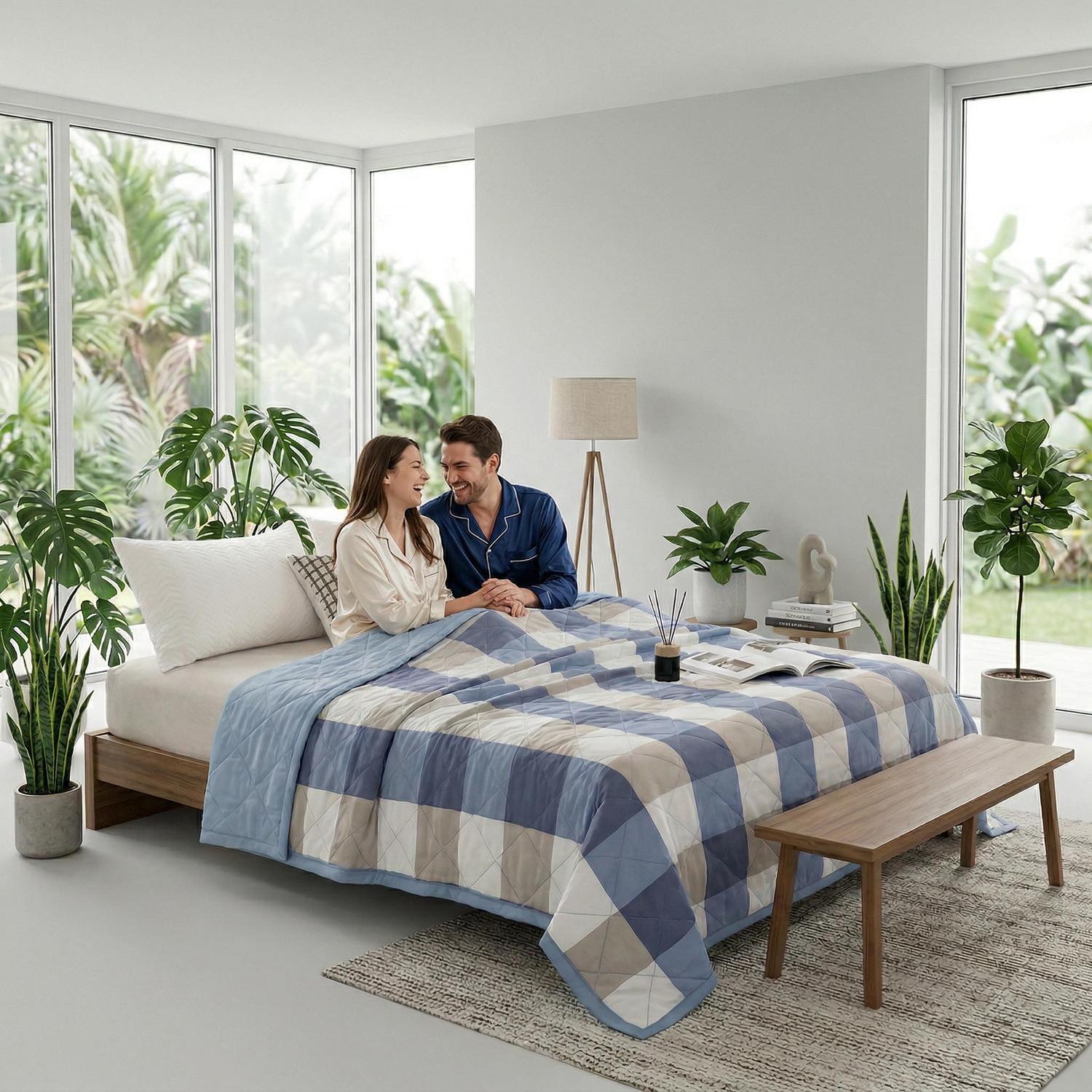 Couple on a bed with a blue and beige plaid lightweight summer quilt, surrounded by plants.