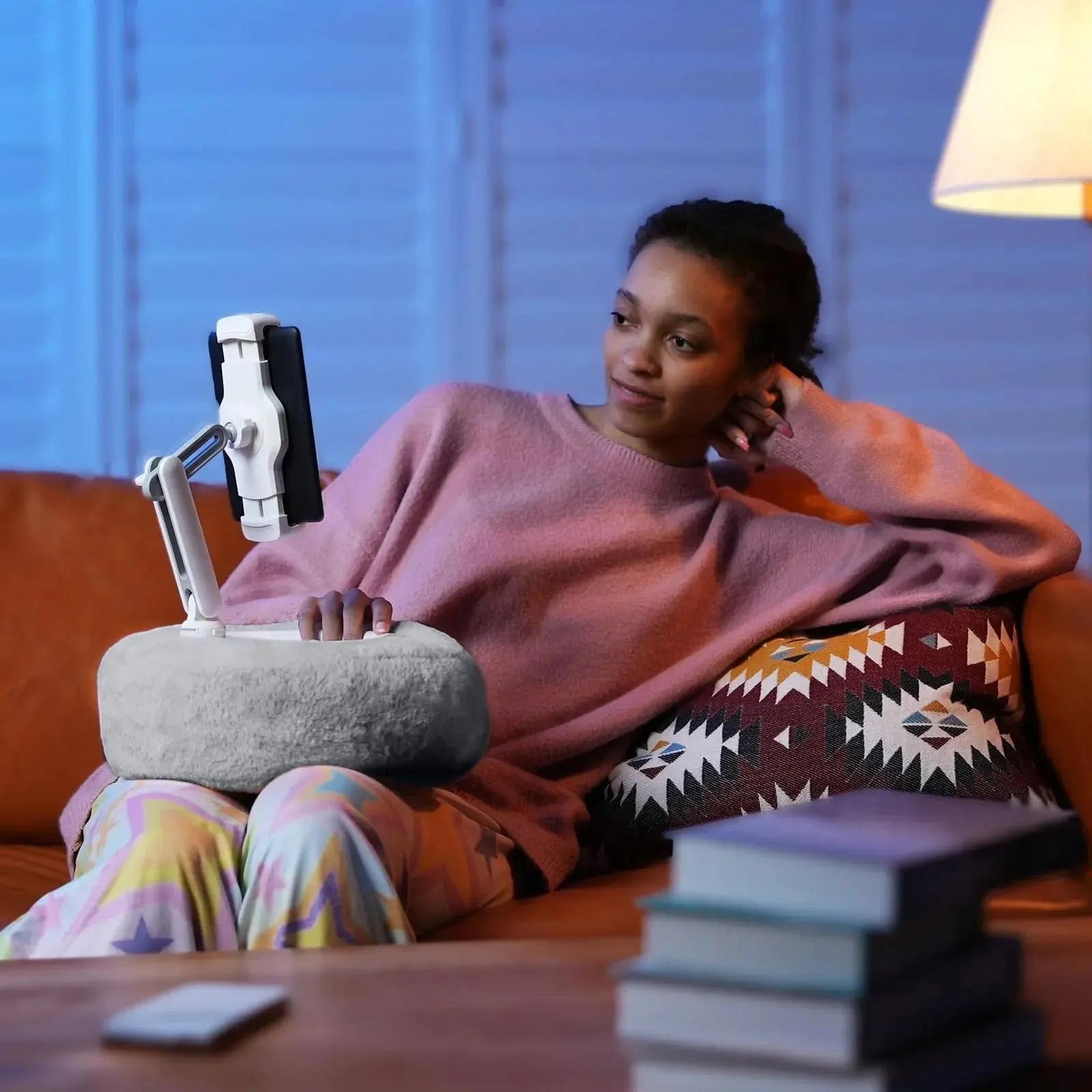 Woman relaxing on a sofa, using a gray plush adjustable device pillow stand to hold her tablet.