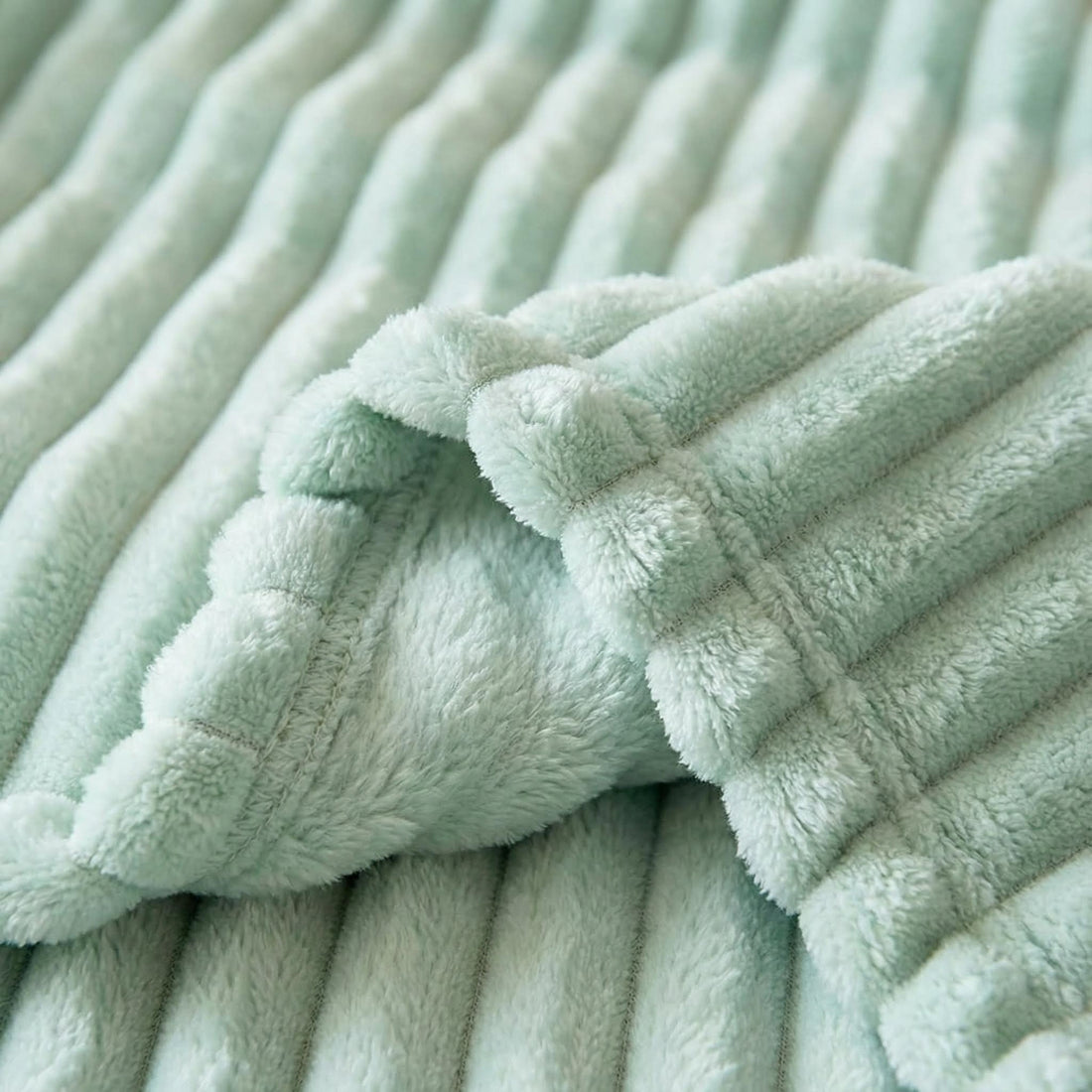 Blonde woman wrapped in a mint green plush faux fur throw blanket with a striped design, holding a mug in a modern bedroom.