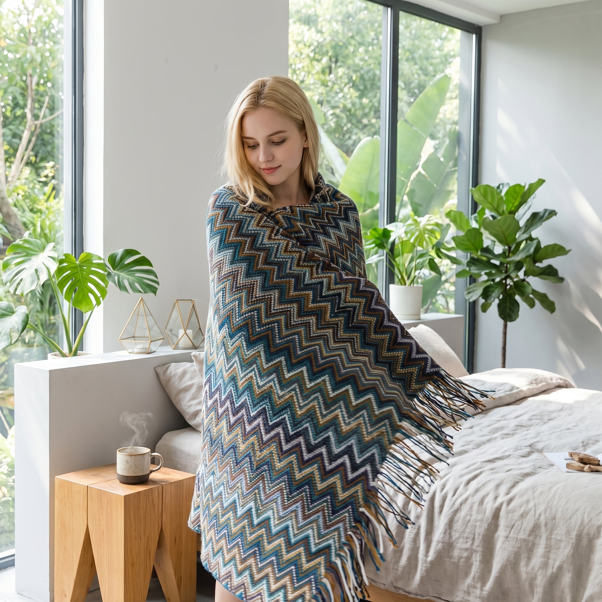 Blonde woman wrapped in a retro bohemian striped knitted blanket with tassels in a bright bedroom.