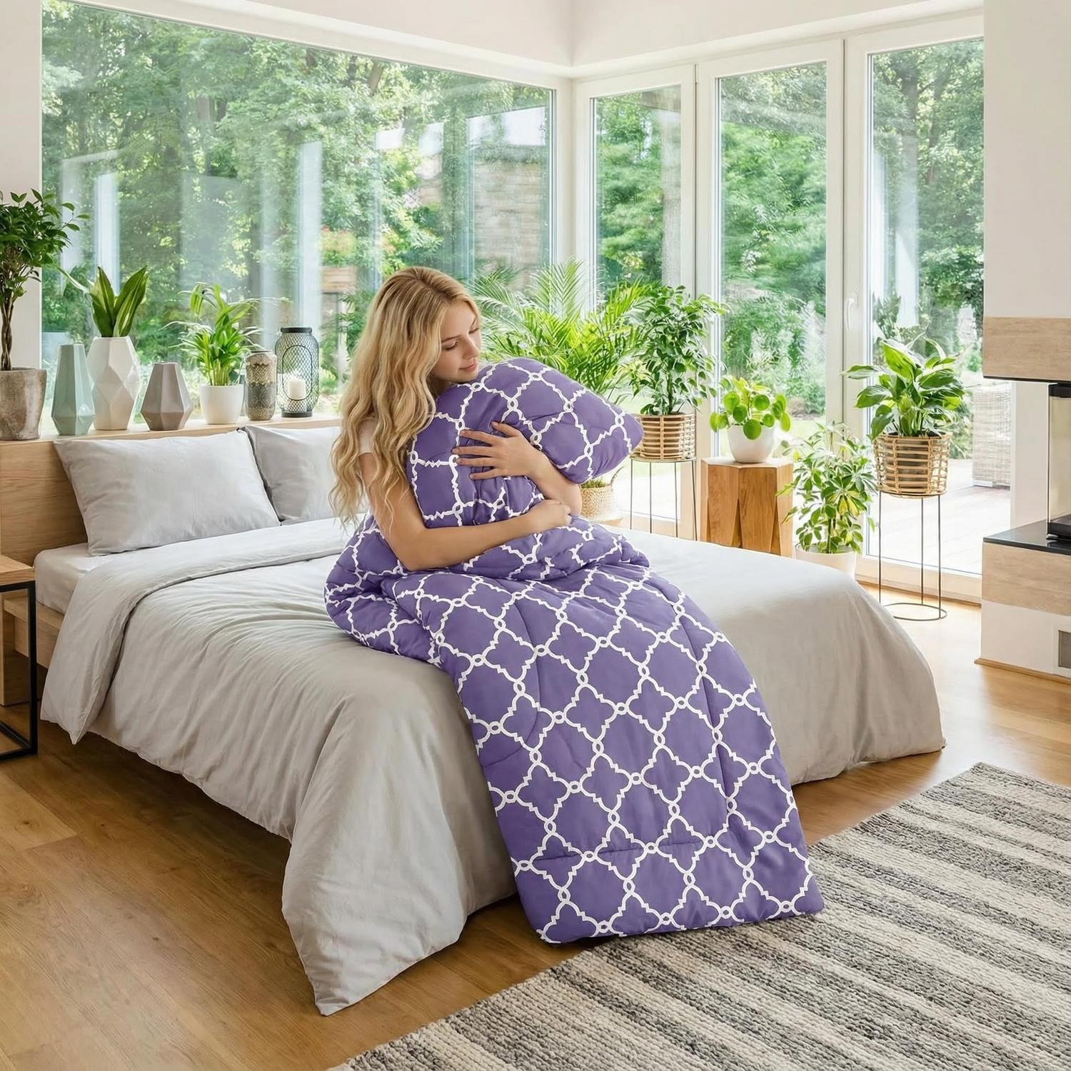 Blonde woman on bed hugging a lavender reversible comforter with white quatrefoil pattern.