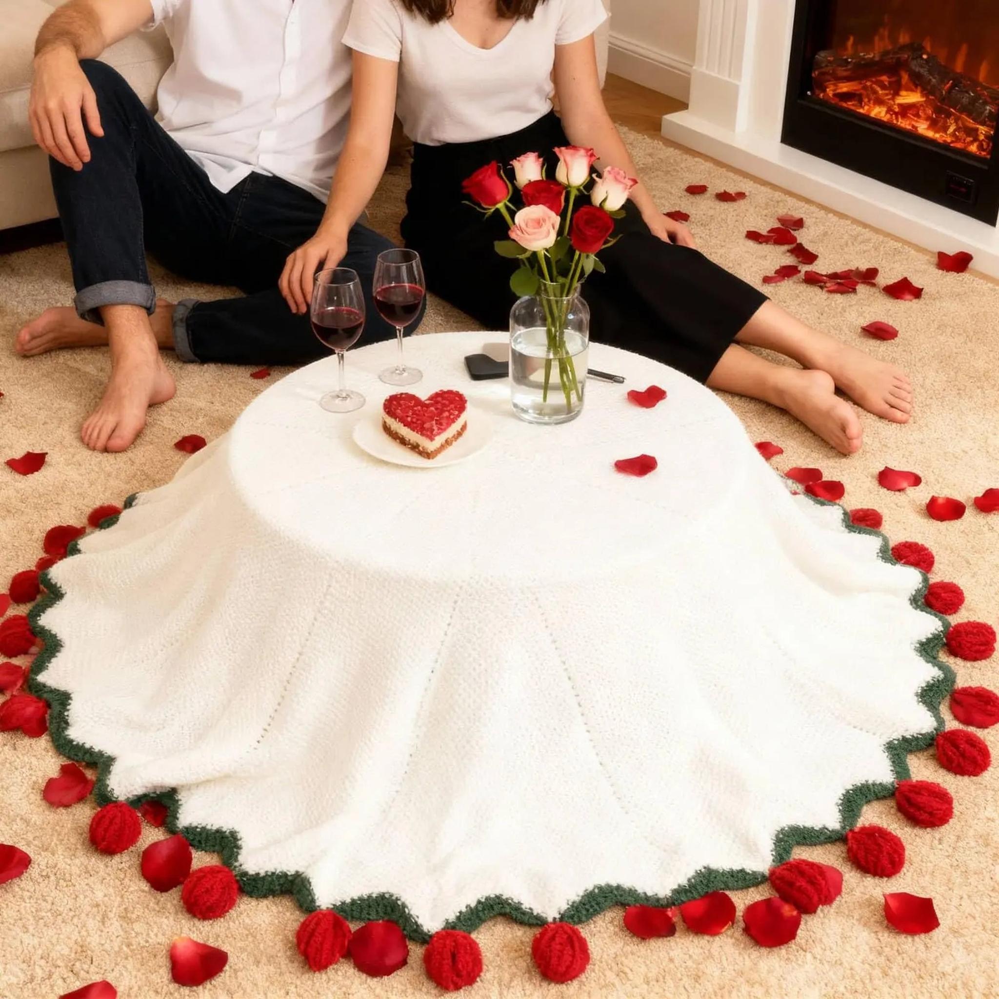 White crochet blanket with rose bouquet fold design, red knitted roses, and green trim, on a carpet with scattered petals.