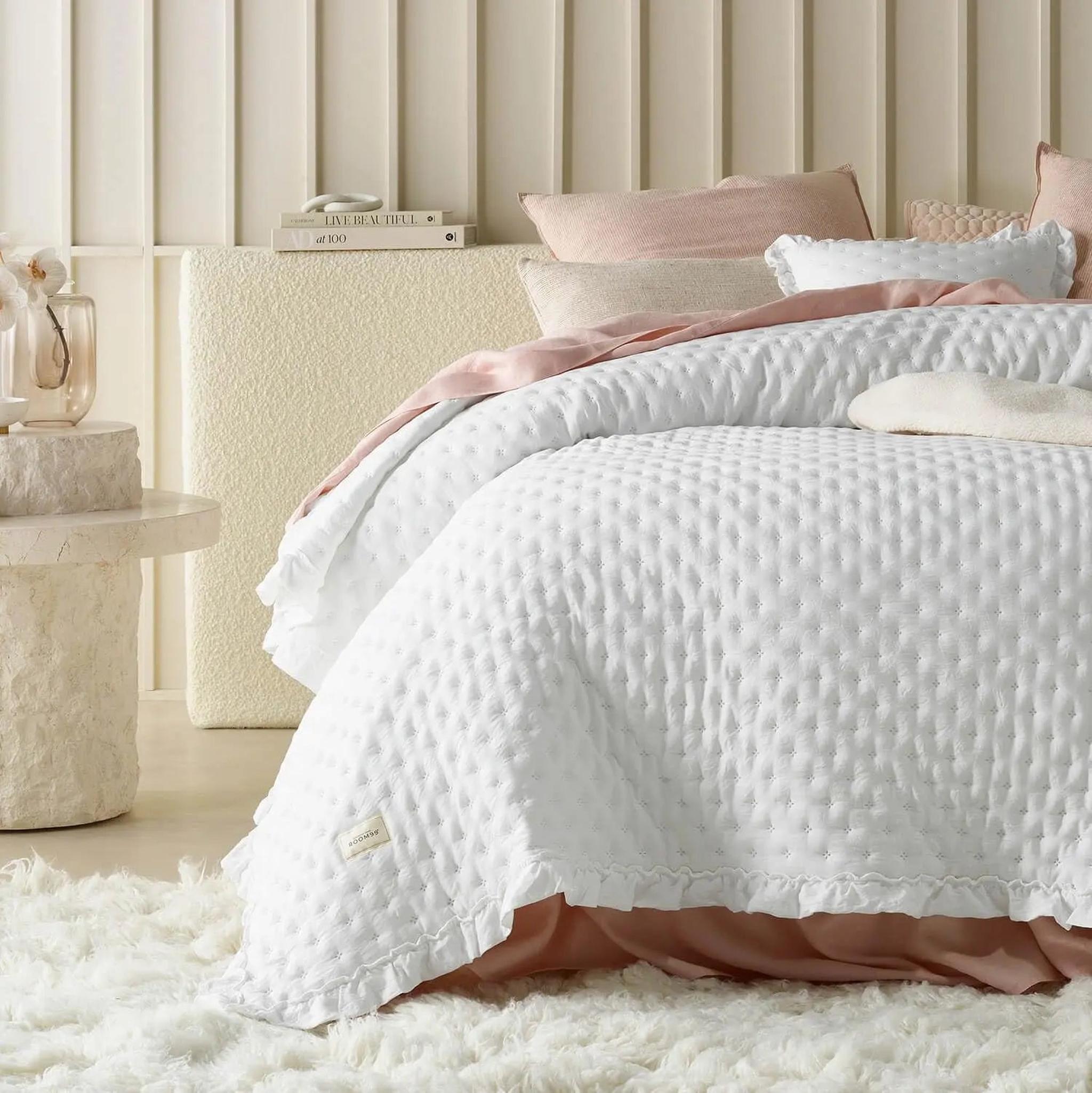 White ruffle mattress cover quilt with scalloped edge, layered over pink bedding on a fluffy rug.