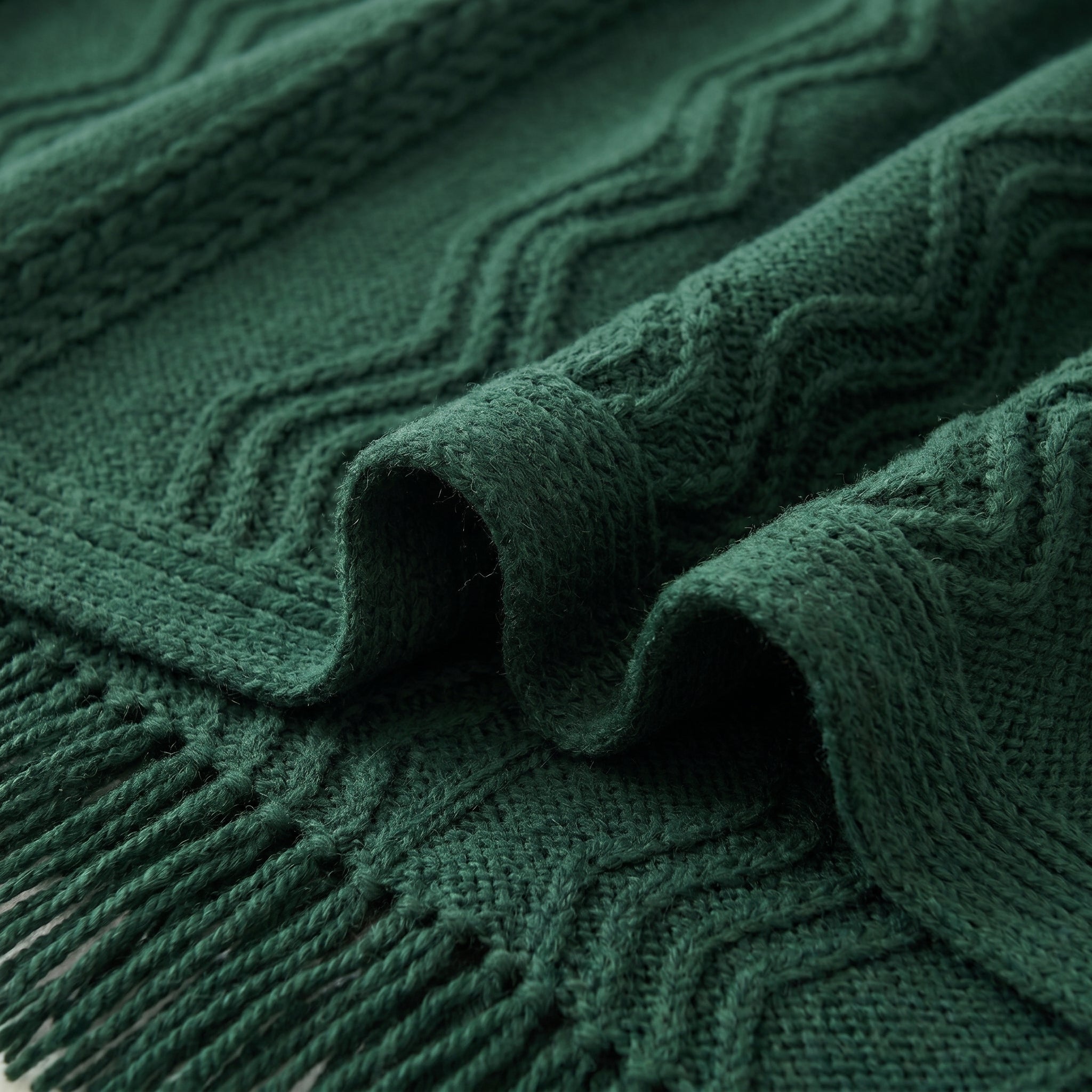 Close-up of a dark green knitted acrylic throw blanket with a wavy plaid pattern and fringe.