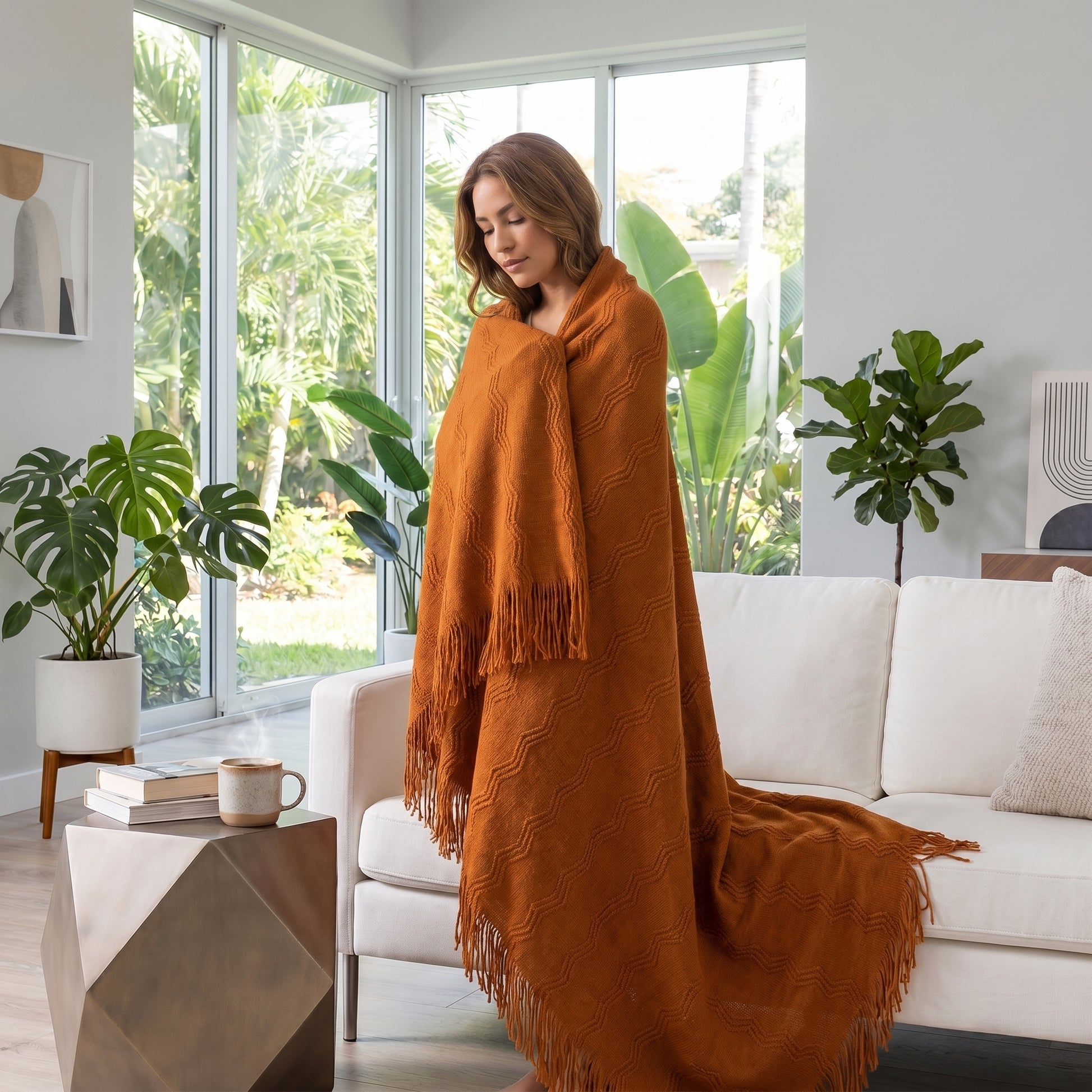 Woman wrapped in a warm, rust-orange knitted throw blanket with fringe, standing in a modern living room.