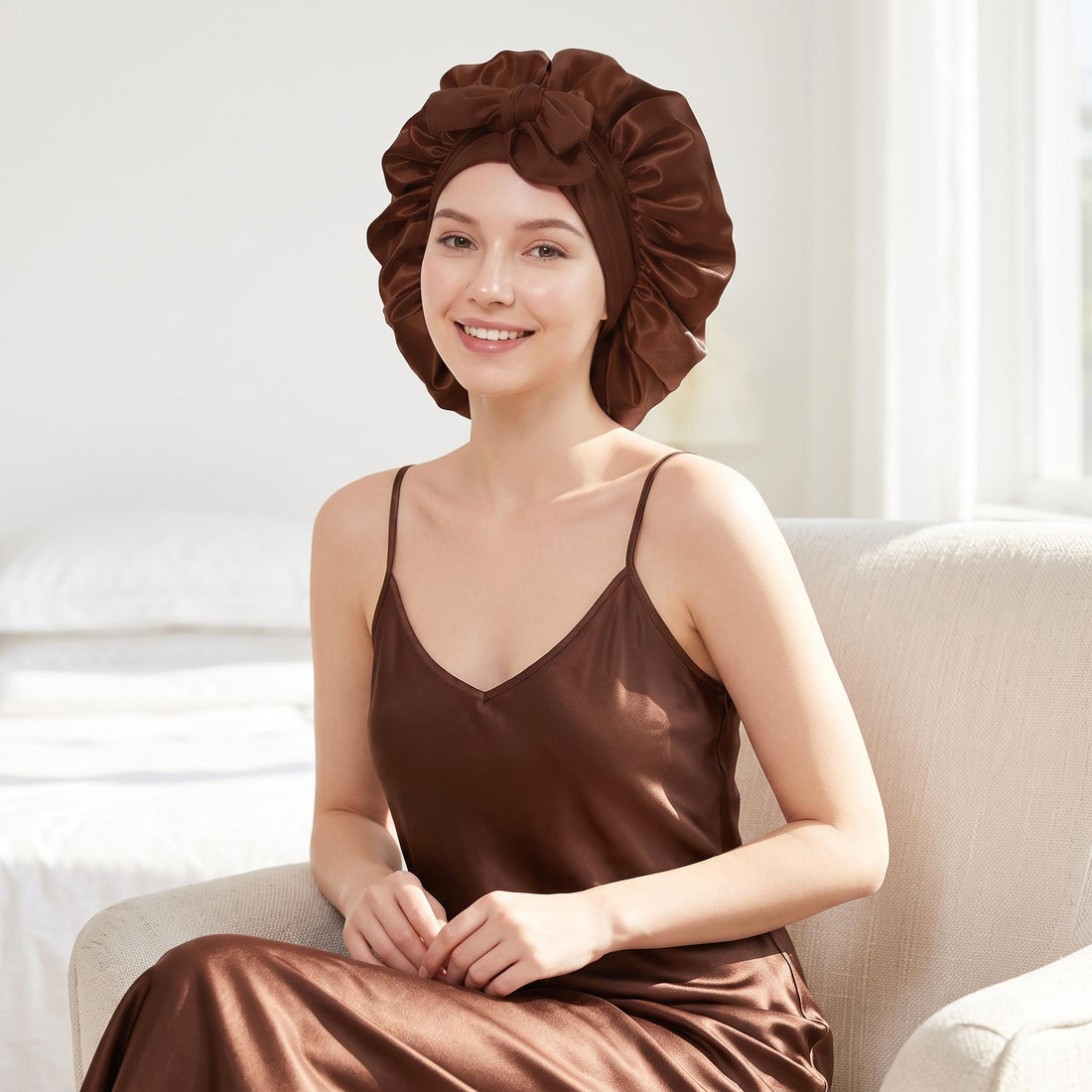 Woman wearing a brown satin silk sleep bonnet with an adjustable tie band while sitting in a chair.