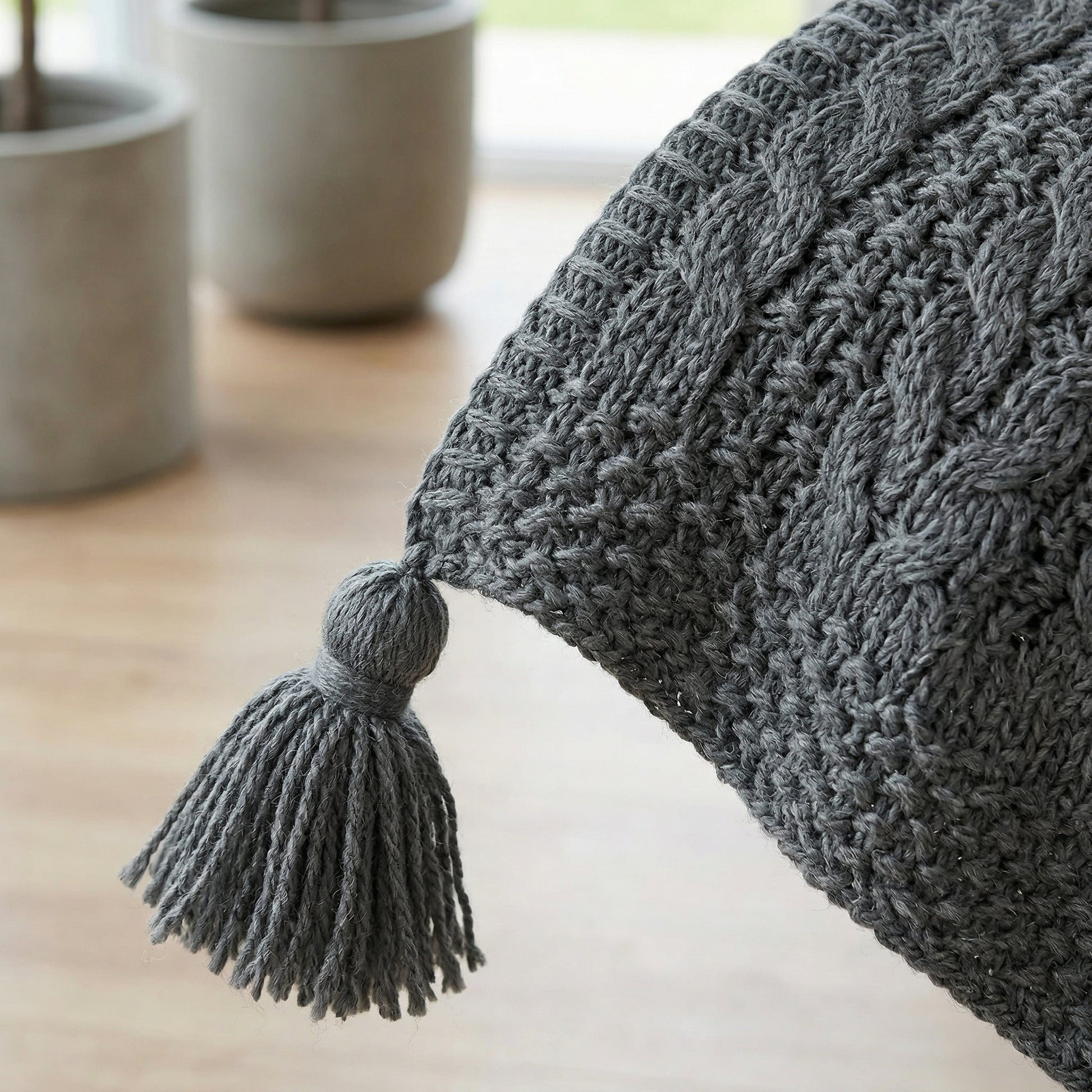 Close-up of a gray knitted acrylic throw blanket with a decorative tassel and cable knit texture.