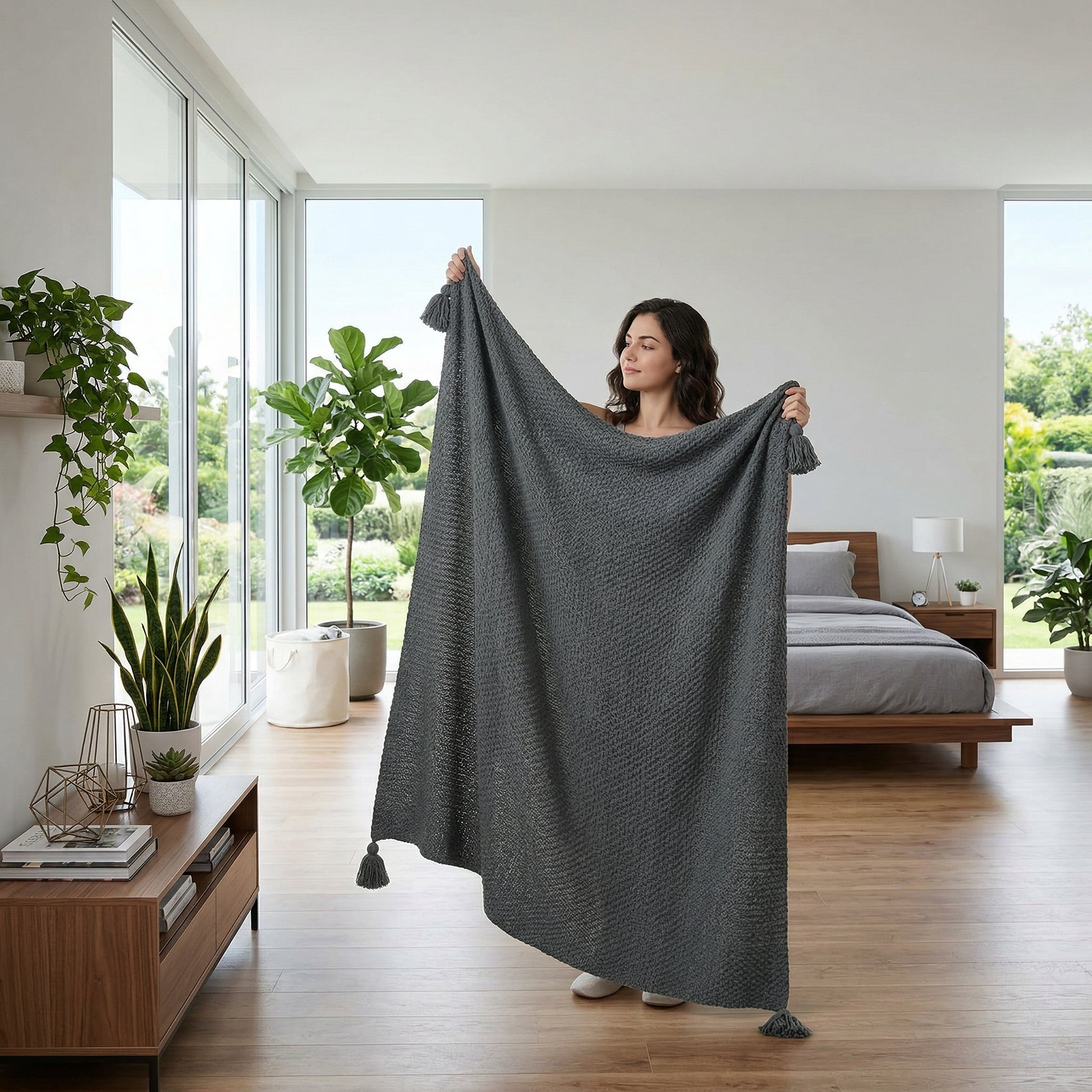Woman holding up a dark gray knitted acrylic throw blanket with tassels in a bright room.