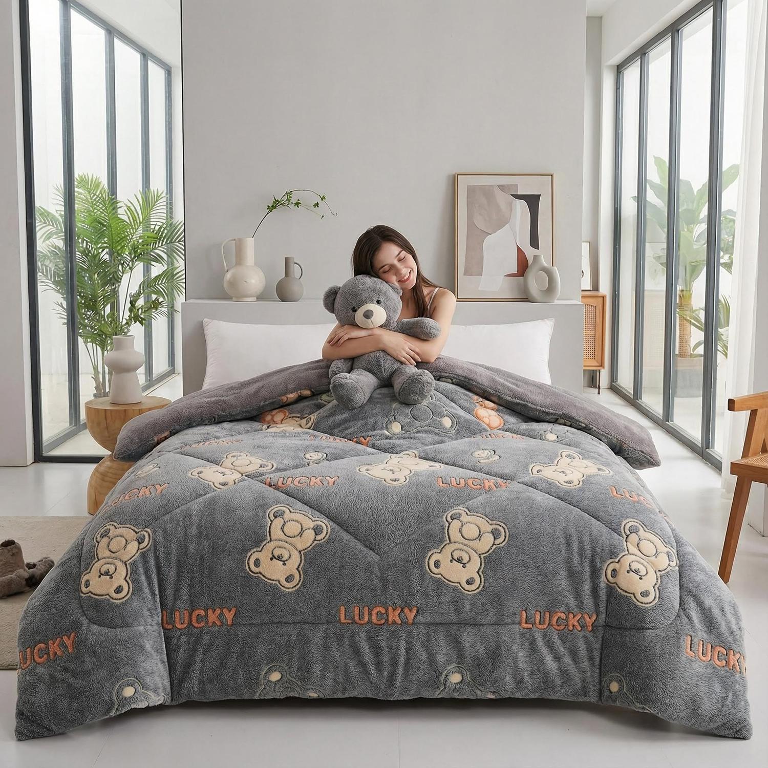 Woman on a bed with a gray velvet quilt featuring a "LUCKY" and teddy bear print, holding a gray teddy bear.