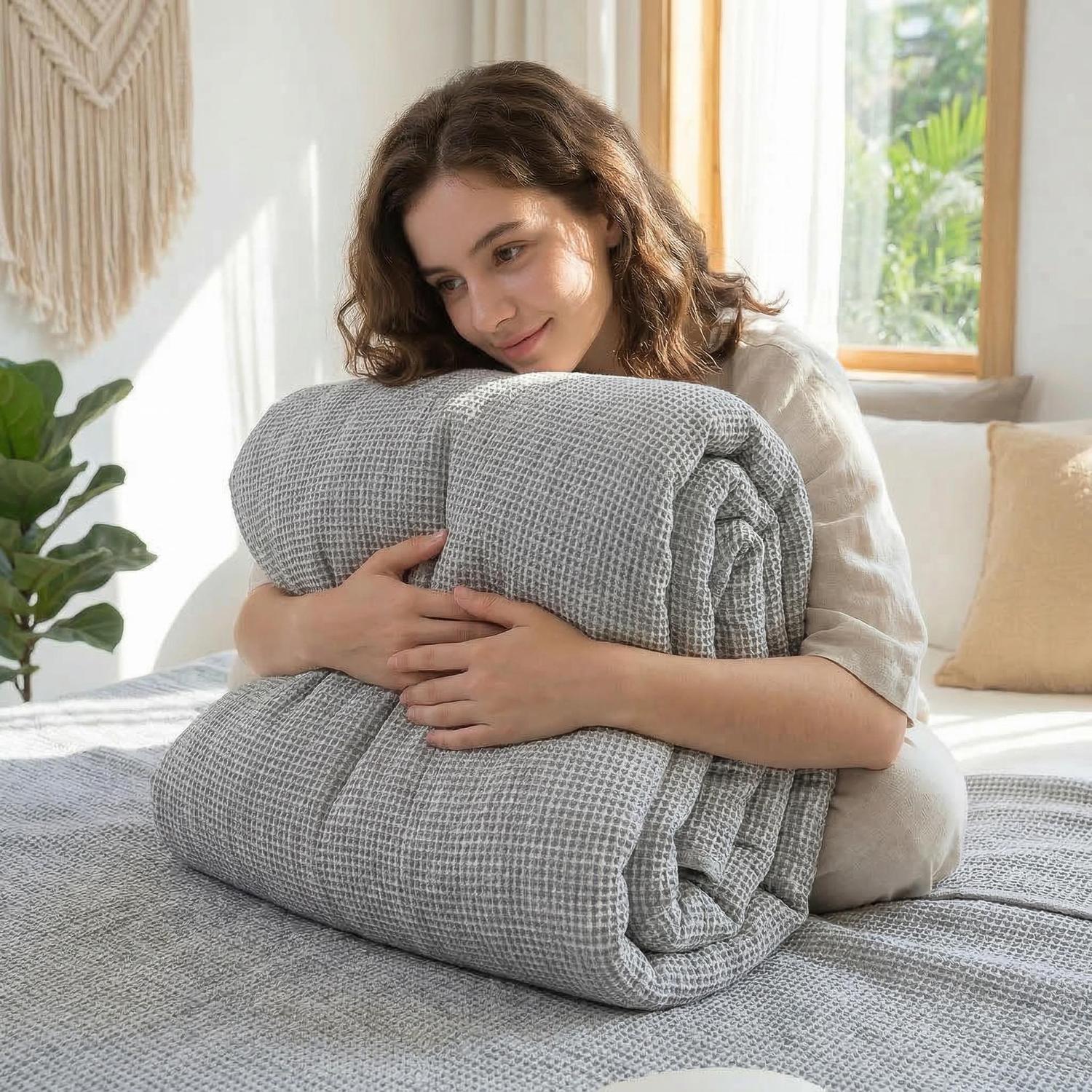 Woman hugging a folded soft gray muslin cotton bedspread with a geometric knitted texture on a bed.