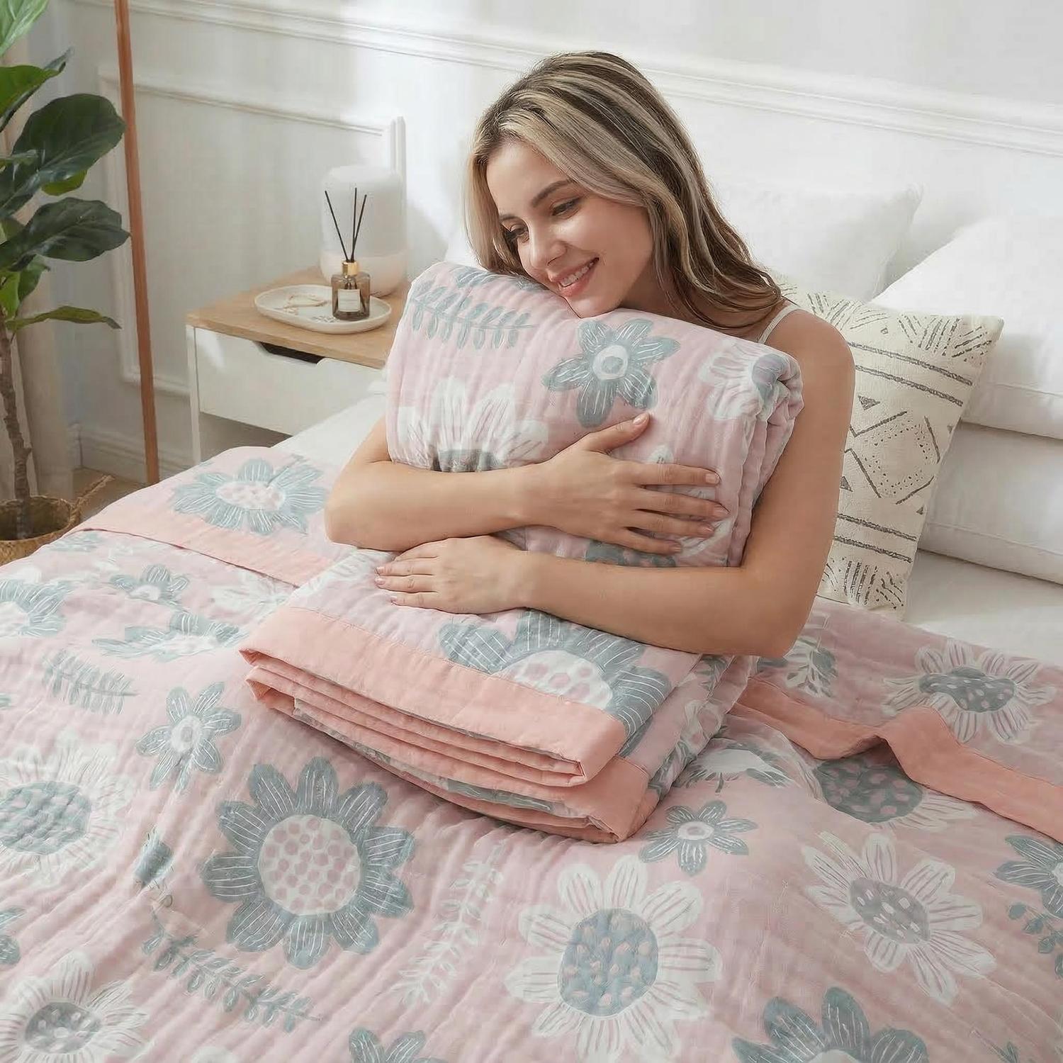 Smiling woman hugging a soft pink cotton bedspread with gray and white floral patterns on a bed.