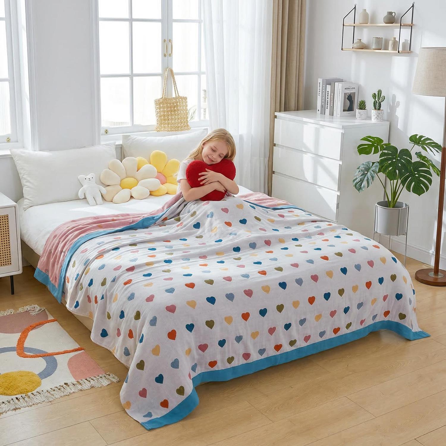 Girl hugging a red heart pillow under a white muslin cotton bedspread with colorful heart patterns and a blue border.