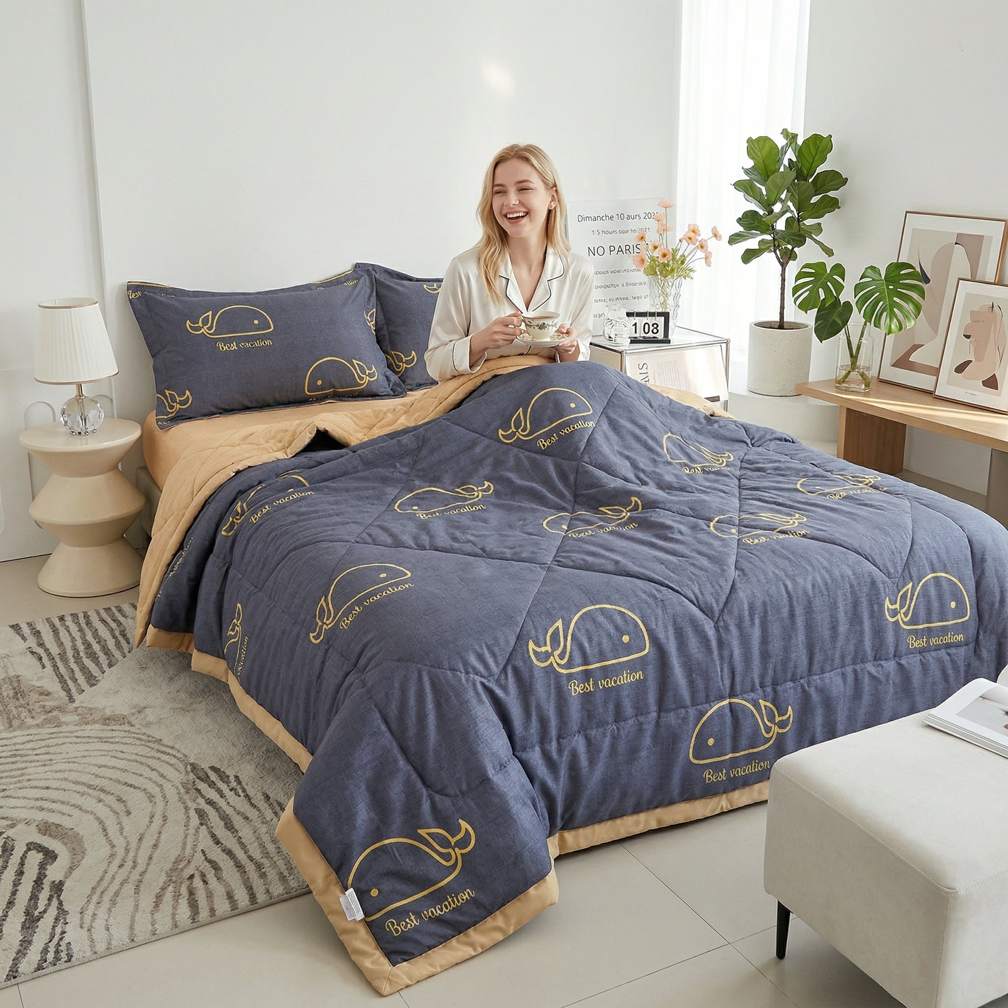 Smiling woman on a bed with a gray and beige Summer Breeze Lightweight Blanket featuring gold whale designs.