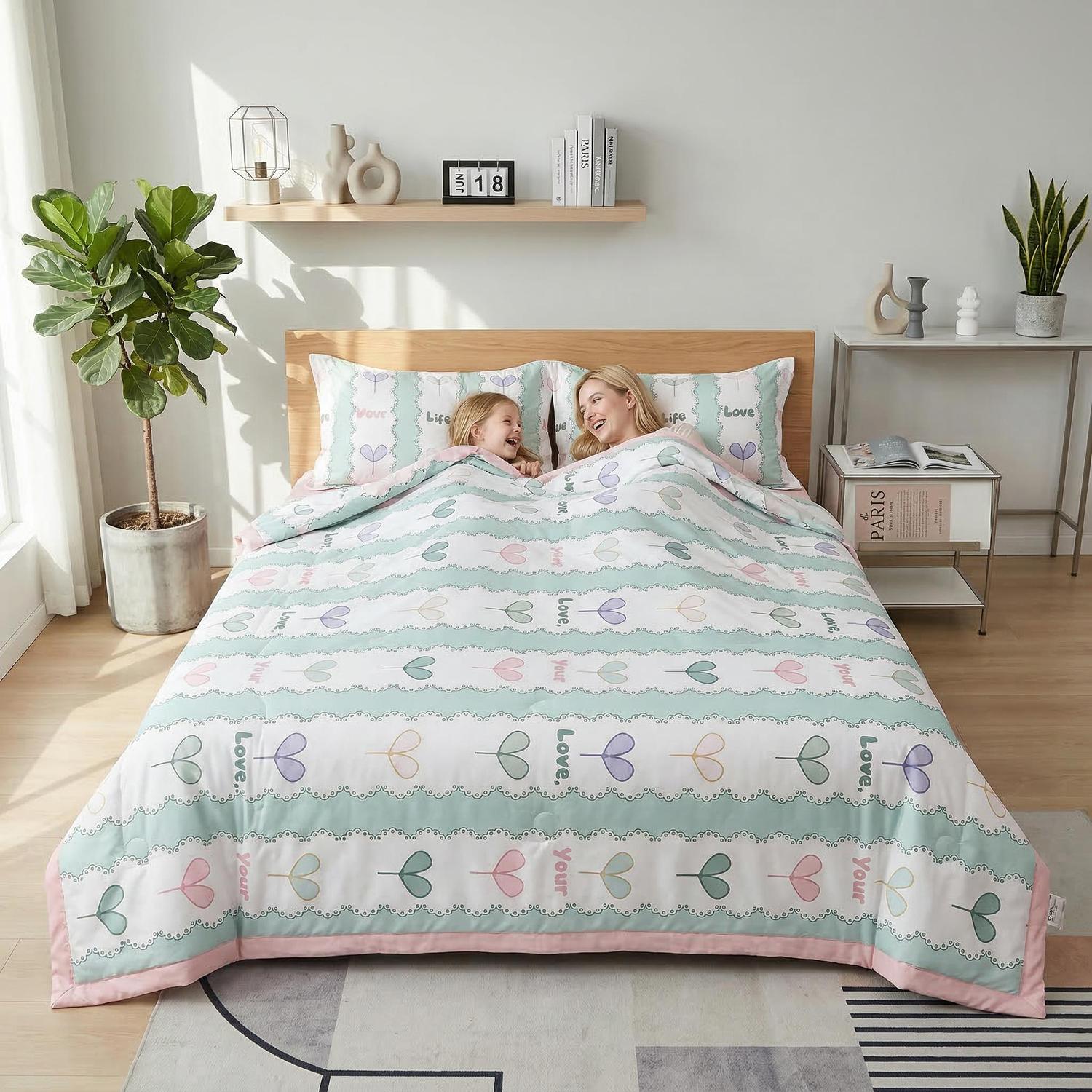 Two people under a white ice silk quilt with pastel heart and "Love" patterns, pink trim.
