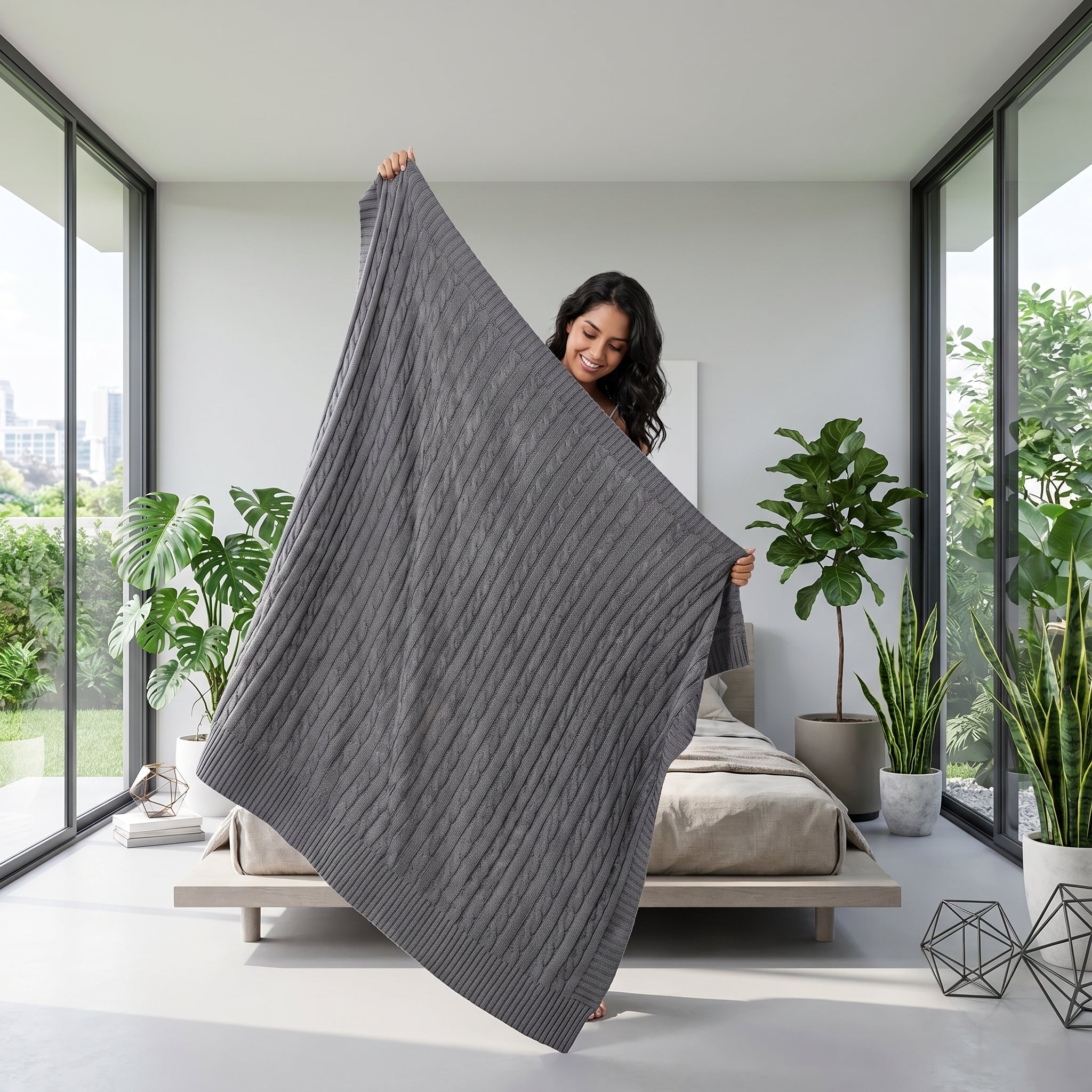 Smiling woman holding up a dark gray textured cable knit cotton throw blanket in a modern bedroom.