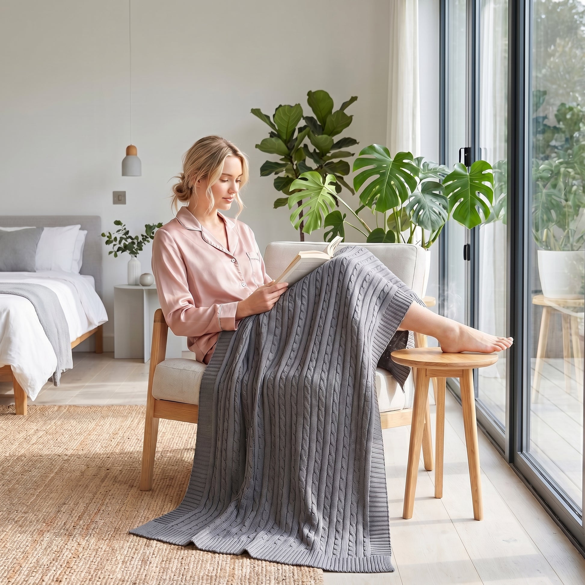Woman in pink pajamas reading, cozy under a gray textured cable knit cotton throw blanket in a bright room.