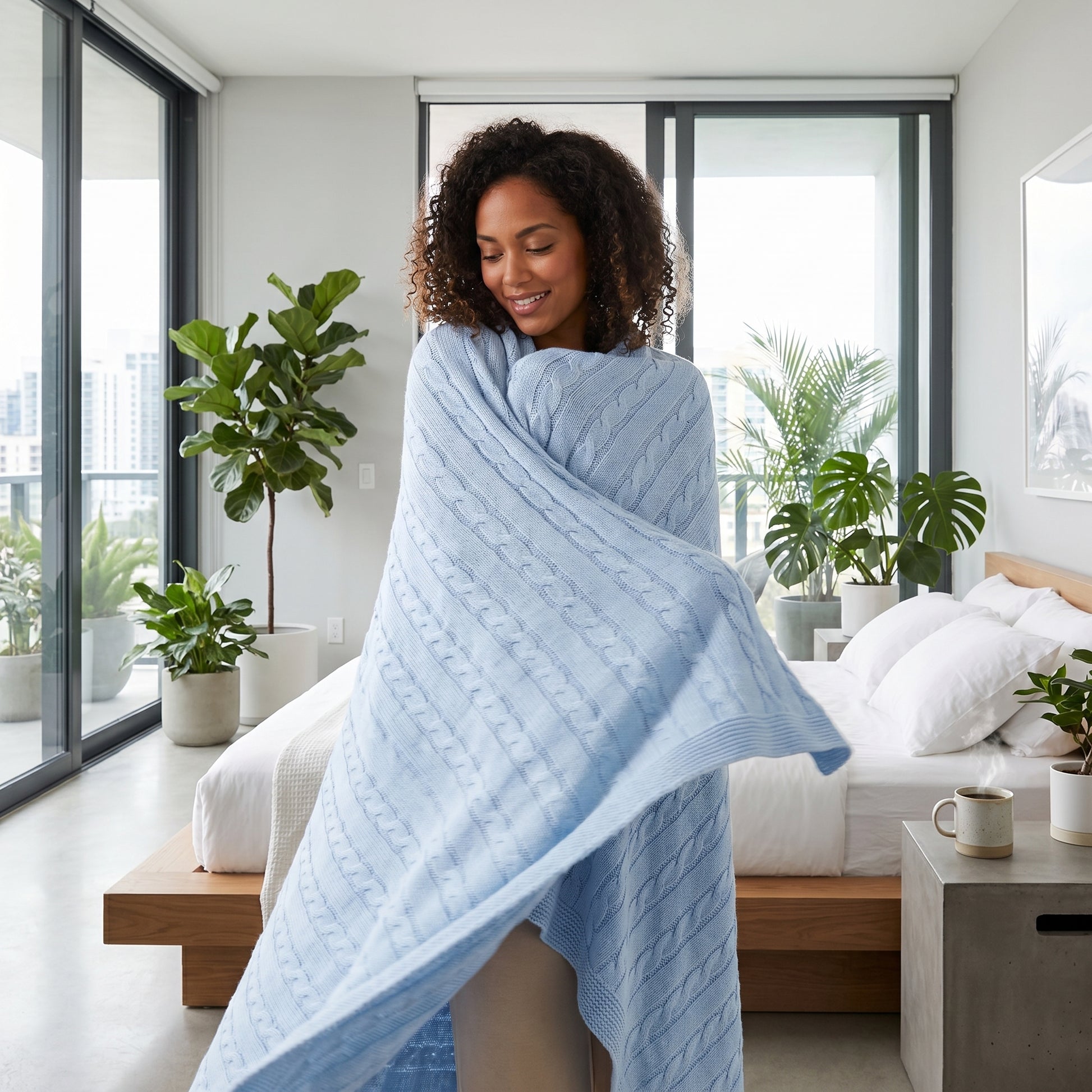 Smiling woman wrapped in a light blue textured cable knit cotton throw blanket in a modern bedroom.