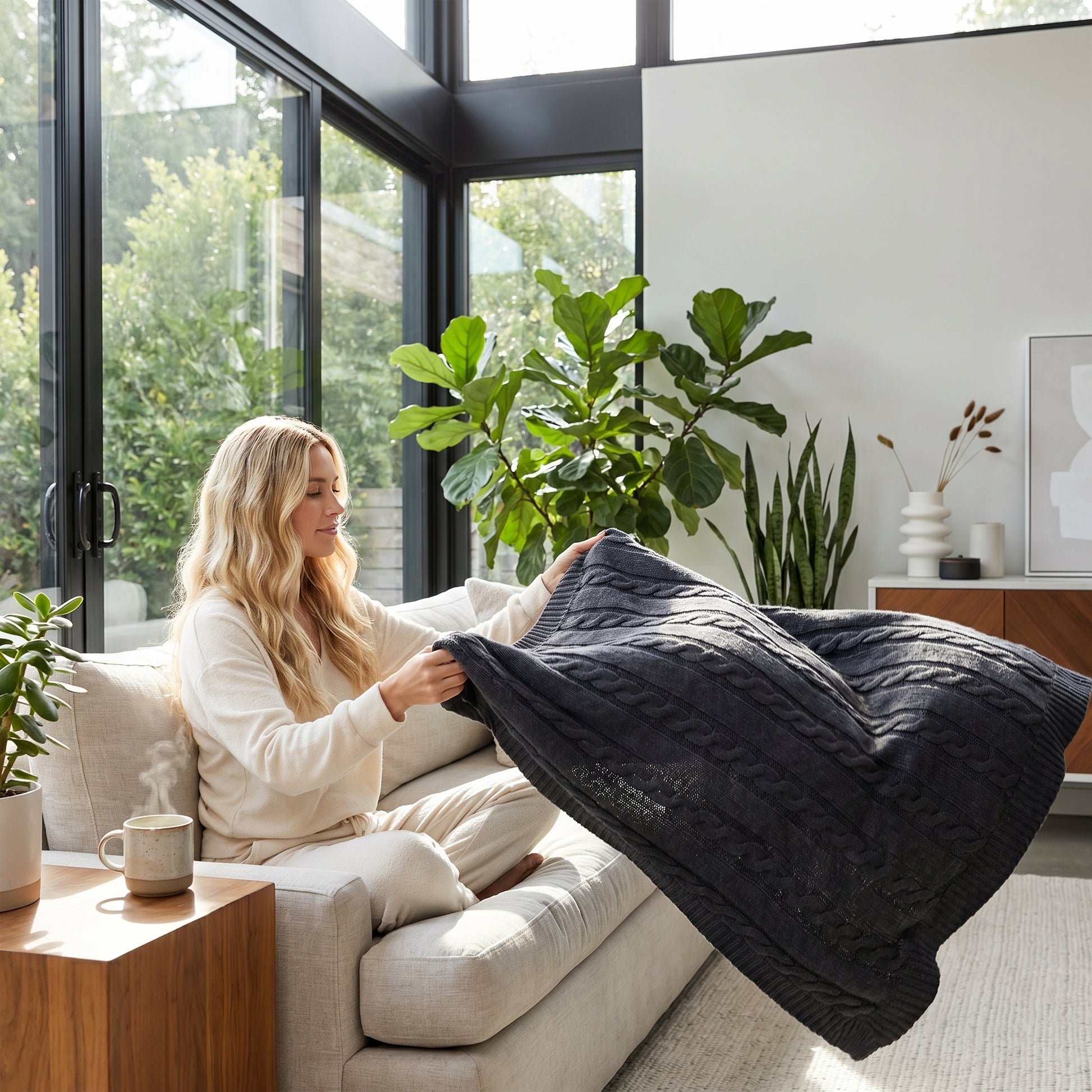 Blonde woman on a sofa holding a dark gray textured cable knit cotton throw blanket in a bright living room.