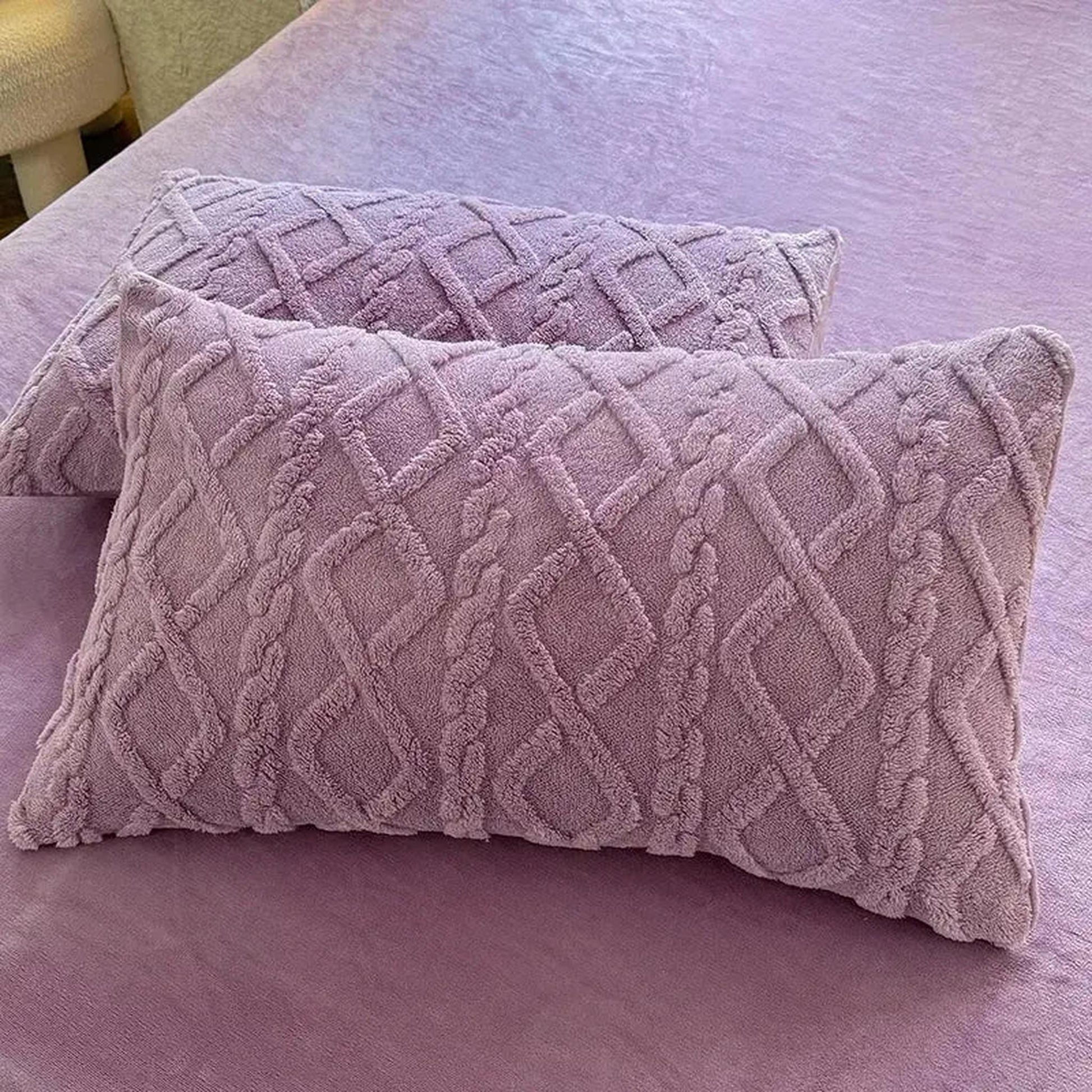 Two soft, plush lavender velvet pillowcases with a textured diamond and braid pattern on a matching bed.