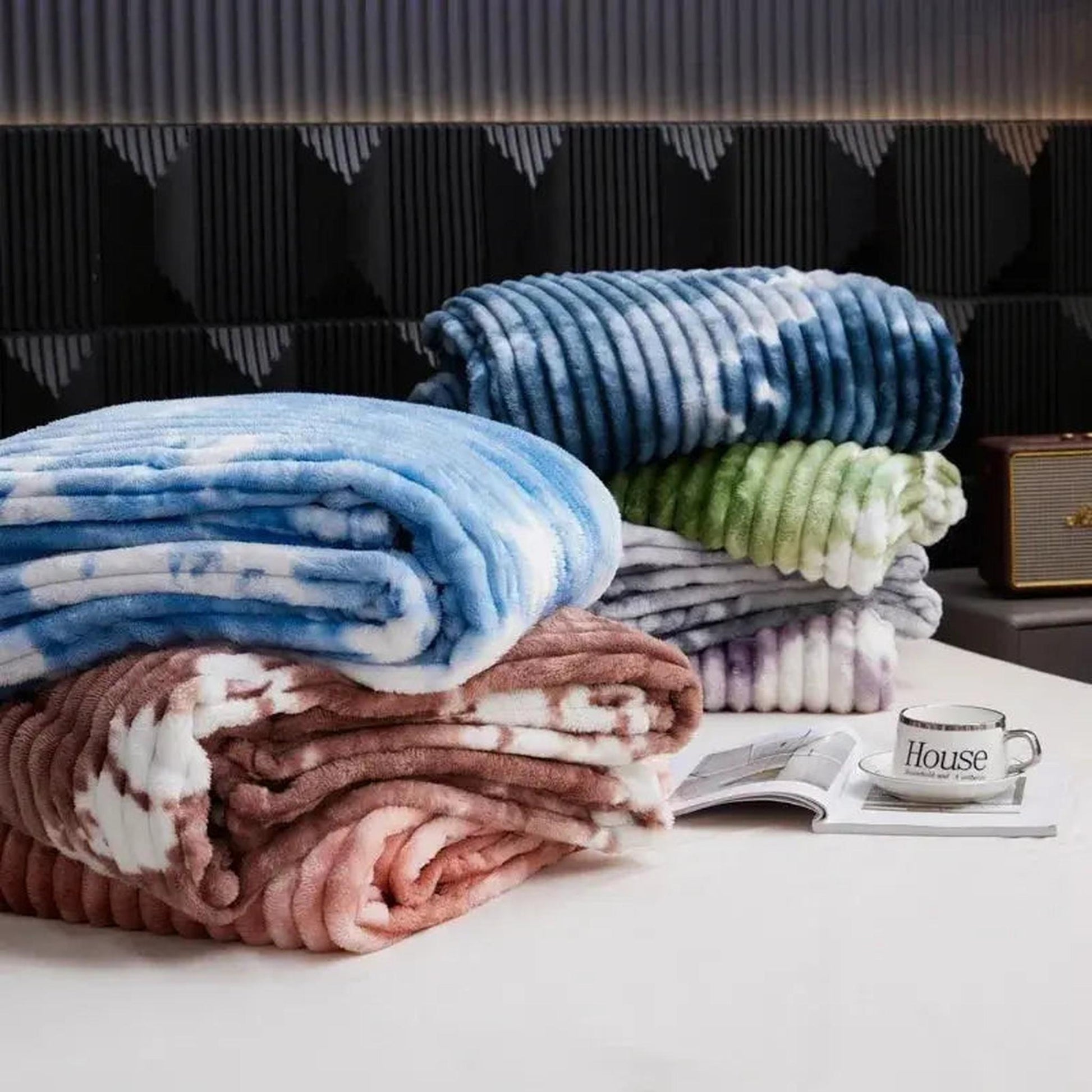 Stack of four colorful tie-dye flannel fleece blankets in blue, green, grey, and pink, on a bed.