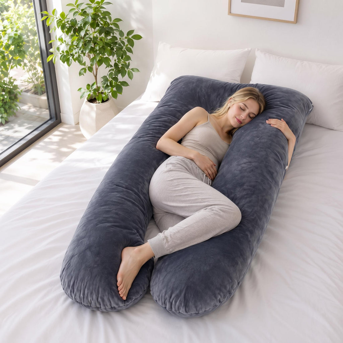 Woman sleeping on a plush, dark grey U-shaped pregnancy pillow on a white bed for full-body support.