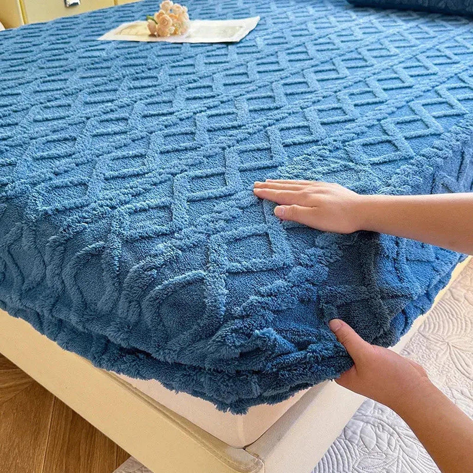Hands adjusting a blue coral fleece fitted bed sheet with a textured ribbed pattern on a mattress.