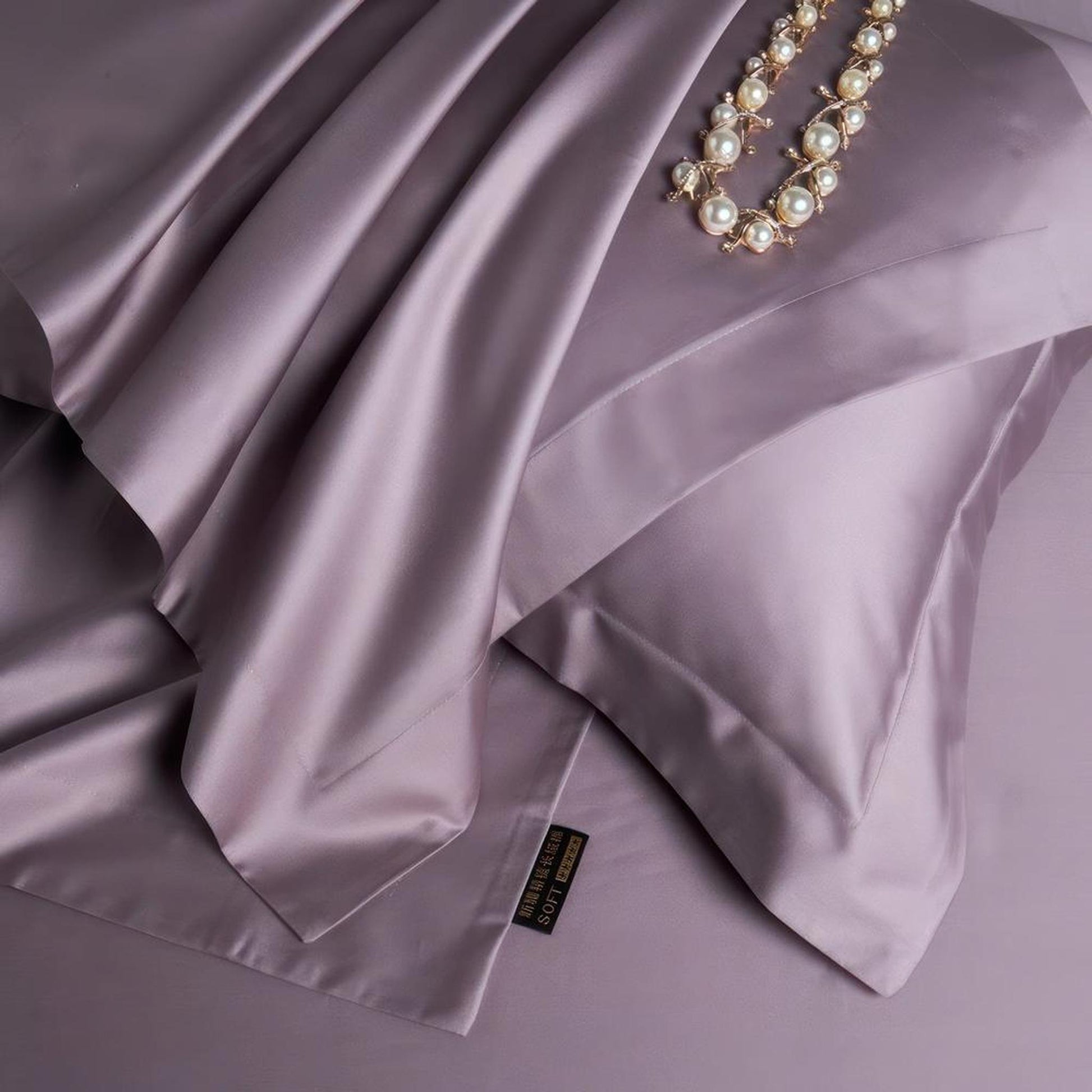 Muted violet silk bedding set with a satin finish, featuring a pillowcase and draped sheet, adorned with a pearl necklace.