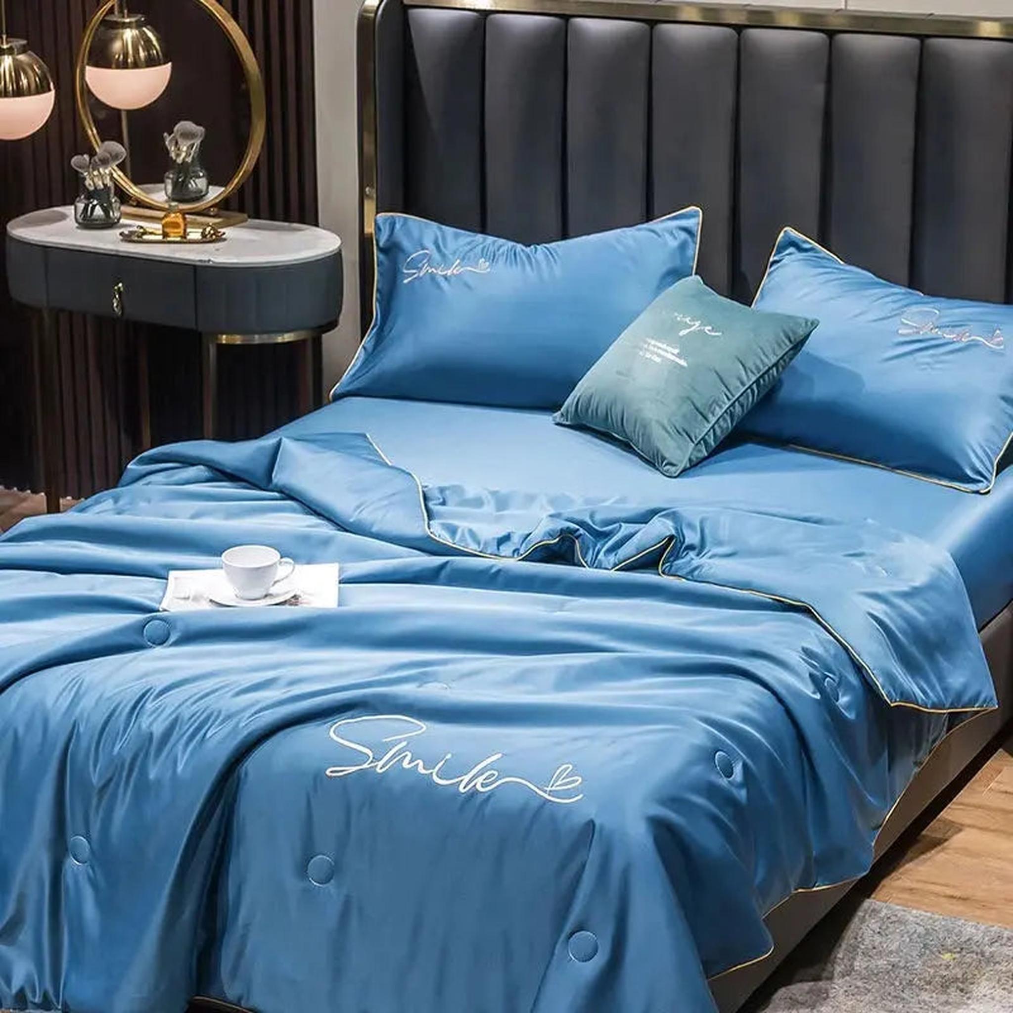 Blue washed silk summer quilt set with embroidered "Smile" design and gold piping on a dark bed.