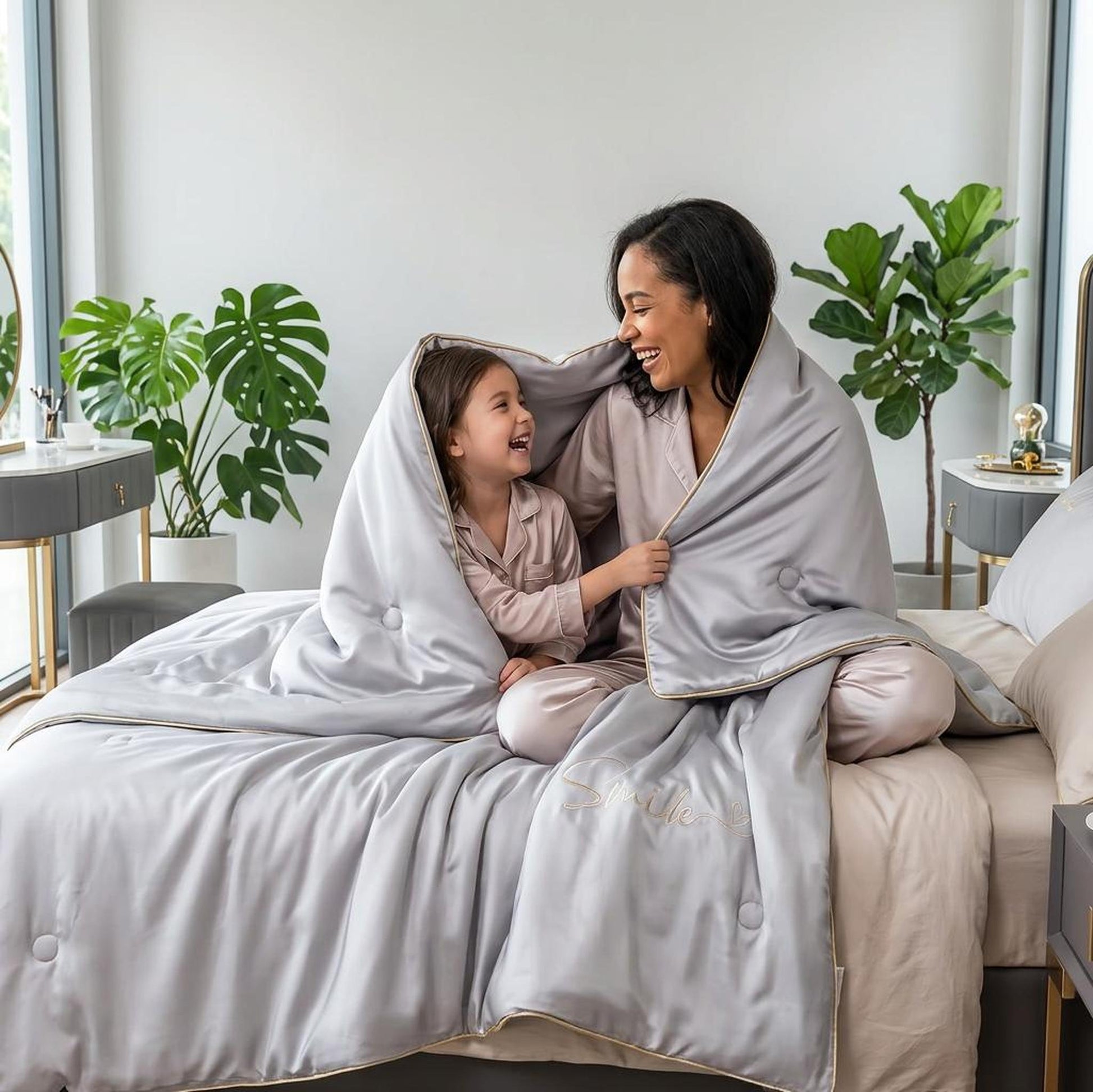 Mother and child laughing under a light gray washed silk quilt with subtle embroidery and gold trim.