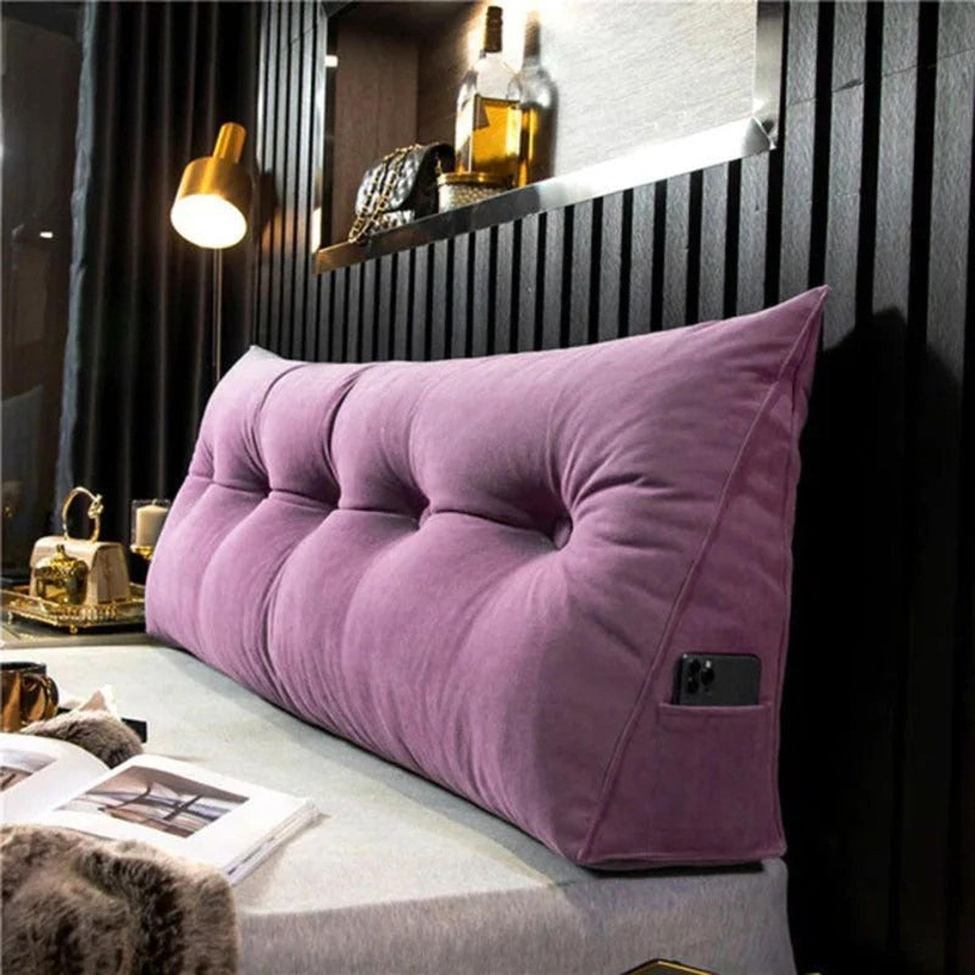 Purple tufted wedge pillow with a side pocket holding a phone, on a bed.
