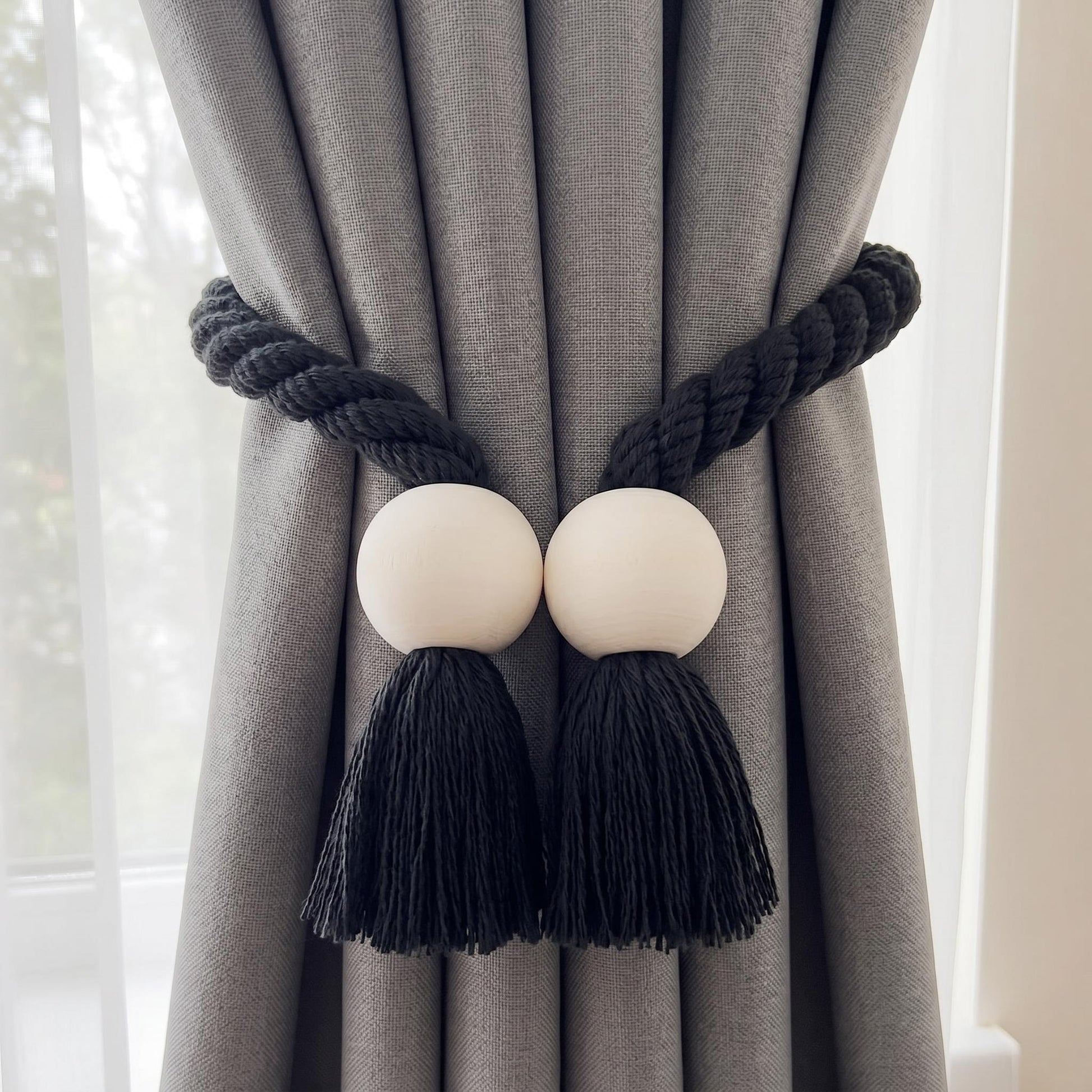 Black braided rope curtain tieback with wooden beads and tassels on a grey curtain.